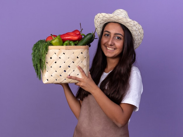 エプロンと夏の帽子の若い庭師の女の子は、紫色の壁の上に元気に立って笑顔で幸せそうな顔と野菜でいっぱいの木枠を保持しています