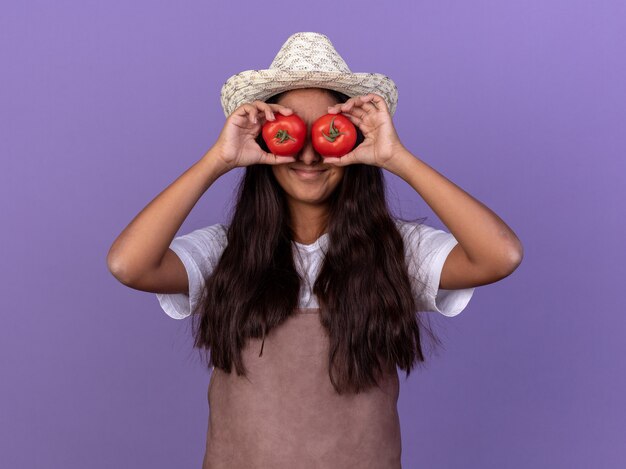 Young gardener girl in apron and summer hat holding fresh tomatoes covering eyes smiling with happy face standing over purple wall