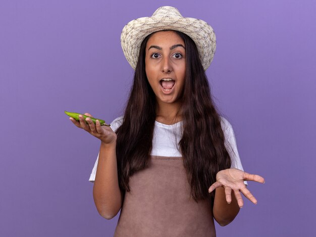 Young gardener girl in apron and summer hat holding green chili pepper  amazed and surprised standing over purple wall