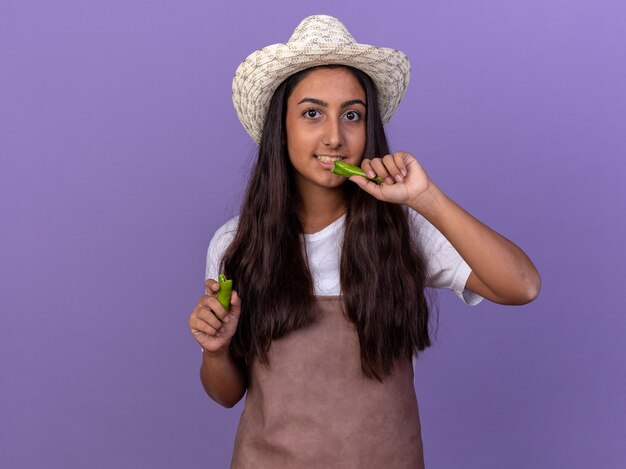 Young gardener girl in apron and summer hat holding green chili pepper going to bite it smiling confused standing over purple wall