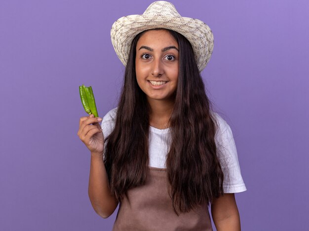 Young gardener girl in apron and summer hat holding green chili pepper  happy and surprised standing over purple wall