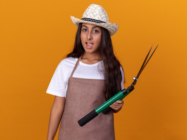 Young gardener girl in apron and summer hat holding hedge clippers  amazed and surprised standing over orange wall