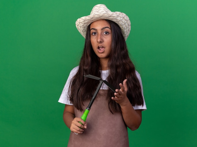 Free photo young gardener girl in apron and summer hat holding mattock  surprised and confused standing over green wall