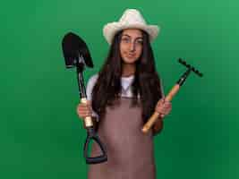 Free photo young gardener girl in apron and summer hat holding mini rake and shovel  with smile on face standing over green wall