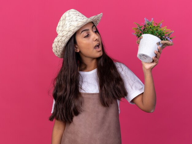 Young gardener girl in apron and summer hat holding potted plant looking at it surprised and amazed standing over pink wall