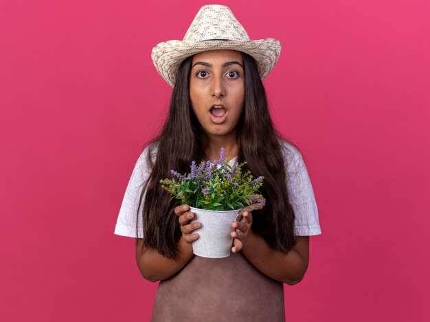 エプロンと鉢植えの植物を保持している夏の帽子の若い庭師の女の子は、ピンクの壁の上に立って驚いて驚いた