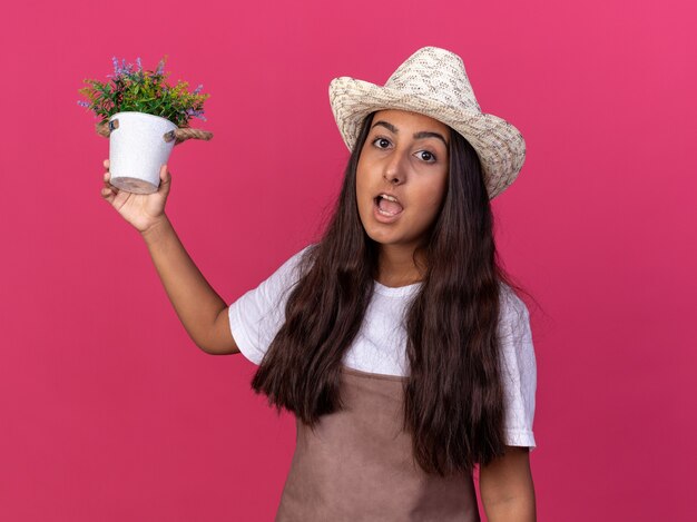 Giovane ragazza del giardiniere in grembiule e cappello estivo che tiene pianta in vaso sorpreso in piedi sopra la parete rosa