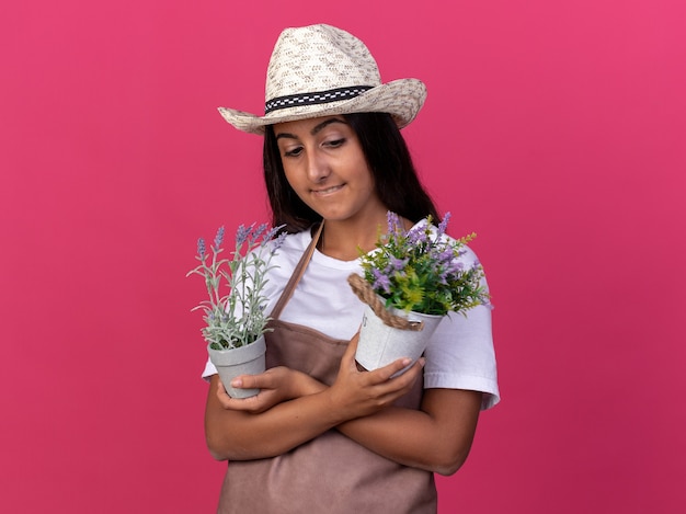 Young gardener girl in apron and summer hat holding potted plants looking confident smiling with smile on face standing over pink wall