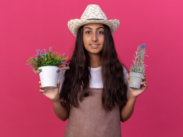 Young gardener girl in apron and summer hat holding potted plants  smiling with happy face standing over pink wall