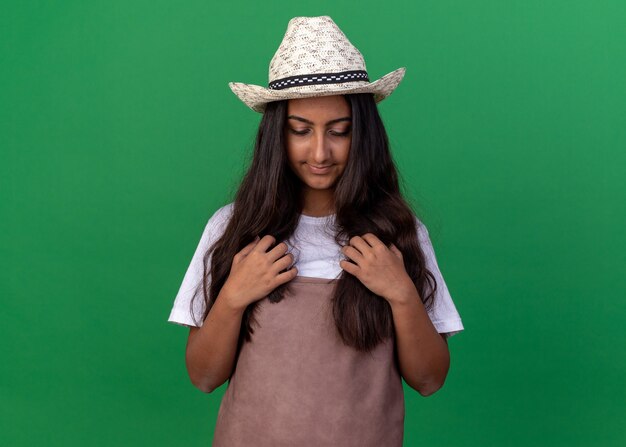 Young gardener girl in apron and summer hat looking down with hands on her chest standing over green wall