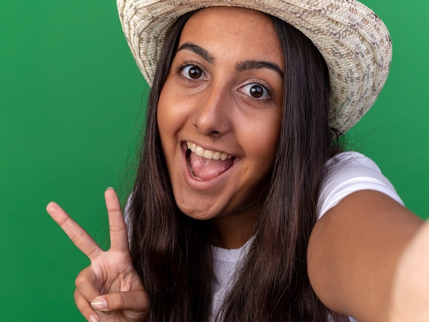 Young gardener girl in apron and summer hat  smiling cheerfully showing v-sign standing over green wall