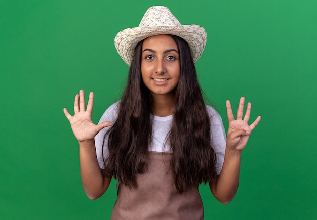 Free photo young gardener girl in apron and summer hat  smiling showing number nine standing over green wall