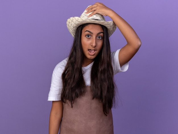 Young gardener girl in apron and summer hat  surprised touching her hat standing over purple wall