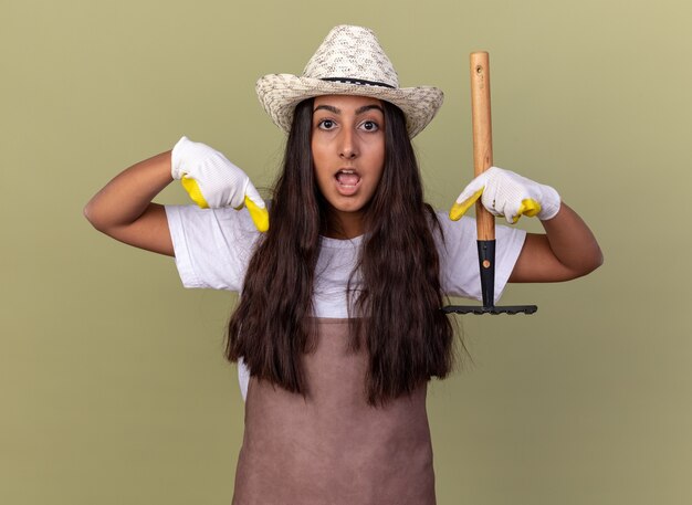 Young gardener girl in apron and summer hat wearing working gloves holding mini rake  surprised pointing with index fingers down standing over green wall