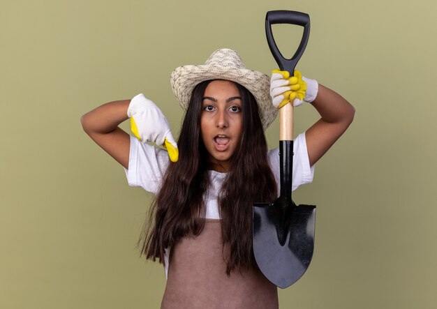 Young gardener girl in apron and summer hat wearing working gloves holding shovel  surprised pointign with index finger down standing over green wall