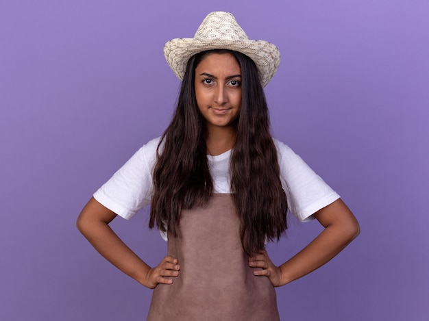 Молодая девушка-садовник в фартуке и летней шляпе со скептической улыбкой на лице стоит над фиолетовой стеной