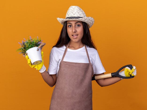 Young gardener girl holding potted plant and shovel with smile on face standing over orange wall