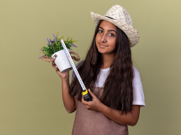 녹색 벽 위에 서있는 얼굴에 미소로 측정 테이프와 측정 테이프를 들고 화분을 들고 앞치마와 여름 모자에 젊은 정원사 소녀