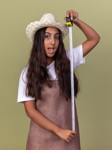 Бесплатное фото Молодая девушка-садовник в фартуке и летней шляпе держит рулетку, счастливая и удивленная, стоя над зеленой стеной