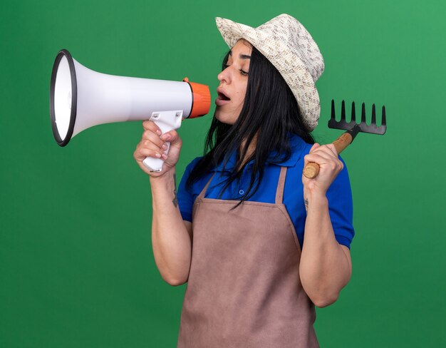 制服を着て帽子をかぶった若い庭師の女の子が緑の壁に隔離されたスピーカーを見下ろして話している熊手を保持している縦断ビュー