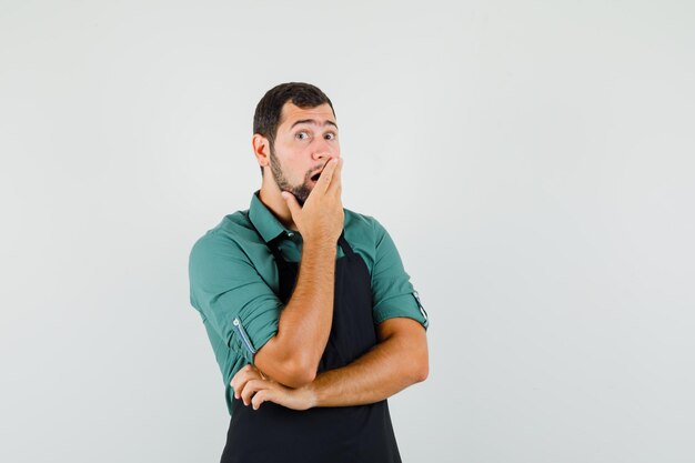 Young gardener holding hand on his mouth in t-shirt,apron and looking surprised , front view.