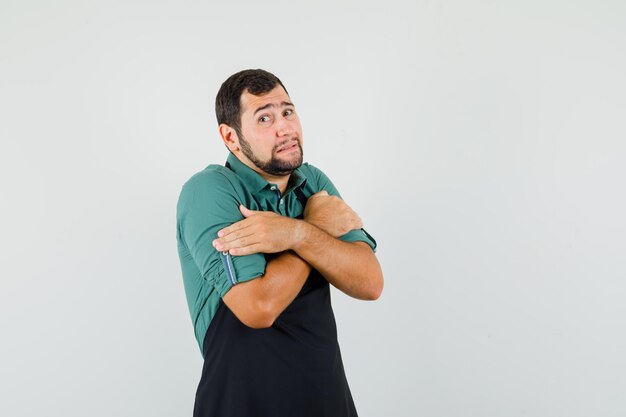 Young gardener hugging himself in t-shirt,apron and looking chilly , front view.