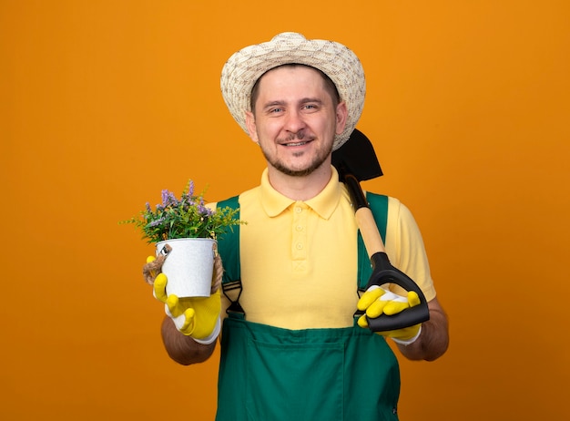 Бесплатное фото Молодой садовник в комбинезоне и шляпе держит лопату и горшечное растение, глядя вперед, улыбаясь со счастливым лицом, стоящим над оранжевой стеной
