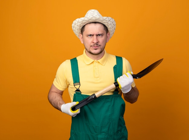 Foto gratuita giovane giardiniere in tuta e cappello tenendo la pala guardando la parte anteriore con la faccia seria in piedi sopra la parete arancione