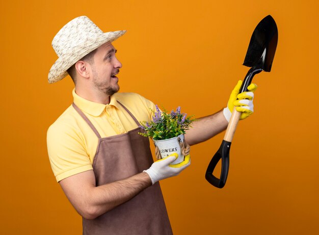 オレンジ色の壁の上に立って笑顔で幸せそうな顔でシャベルを見てシャベルと鉢植えの植物を保持しているジャンプスーツと帽子の若い庭師