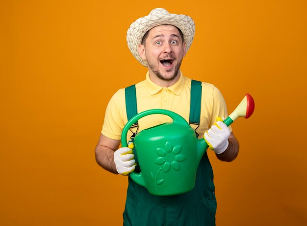 Young gardener in jumpsuit and hat holding watering can looking at front smiling cheerfully standing over orange wall