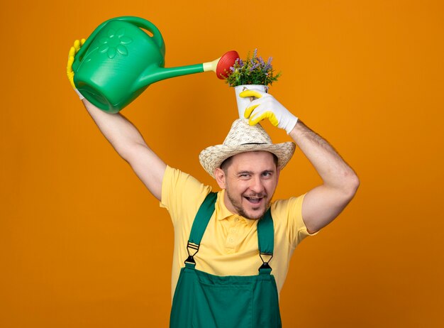 Young gardener in jumpsuit and hat holding watering can and potted plant over his head smiling cheerfully standing over orange wall