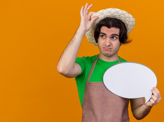 Young gardener man in apron and hat holding blank speech bubble sign looking aside being confused and displeased touching his hat standing over orange wall