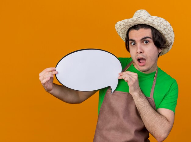 Young gardener man in apron and hat holding blank speech bubble sign looking at camera surprised and amazed standing over orange background