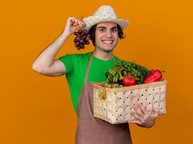 Young gardener man in apron and hat holding crate full of vegetables and bunch of grape looking with happy face smiling broadly standing over orange wall