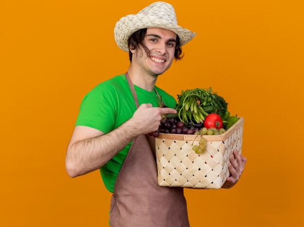 オレンジ色の背景の上に立っている木枠に人差し指で元気に指しているカメラを見て野菜でいっぱいの木枠を保持しているエプロンと帽子の若い庭師の男