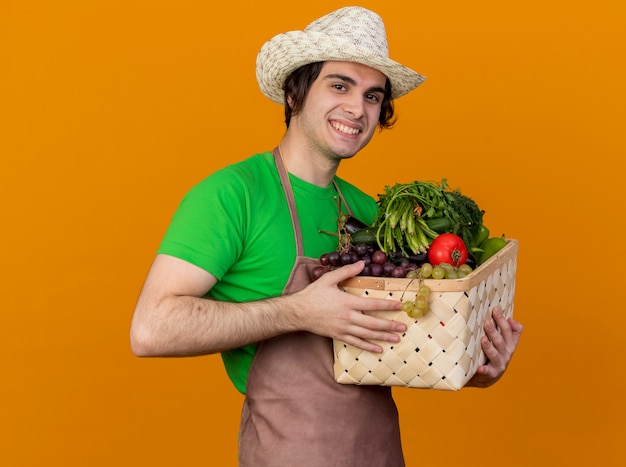 Young gardener man in apron and hat holding crate full of vegetables looking at camera smiling with happy face standing over orange background