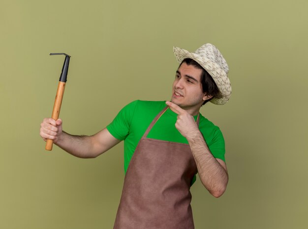 Young gardener man in apron and hat holding mini rake pointing with index finger at it smiling cheerfully standing over light wall