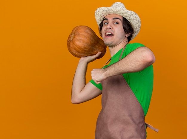 Free photo young gardener man in apron and hat holding pumpkin going to throw int happy and cheerful standing over orange background