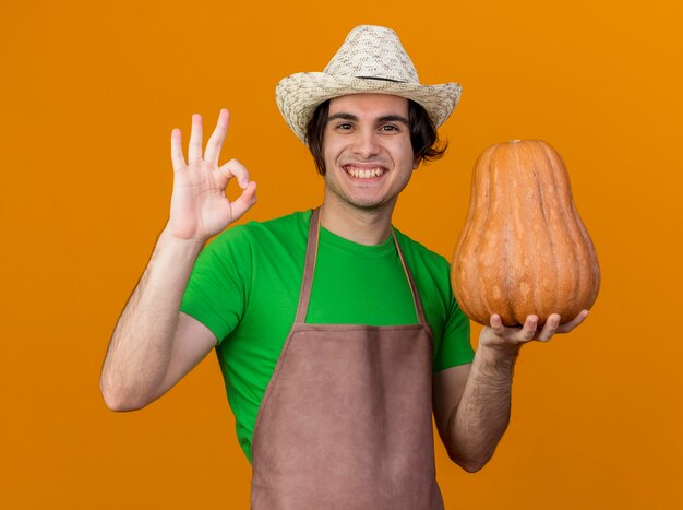 Young gardener man in apron and hat holding pumpkin lookign  smiling cheerfully with happy face showing ok sign standing over orange wall