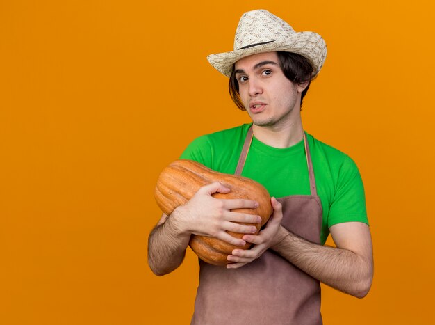 Young gardener man in apron and hat holding pumpkin looking confused standing over orange wall