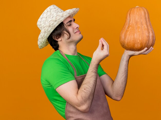 Giovane giardiniere uomo in grembiule e cappello tenendo la zucca guardandolo facendo eccellente cantare con le dita felici e contenti in piedi sopra la parete arancione