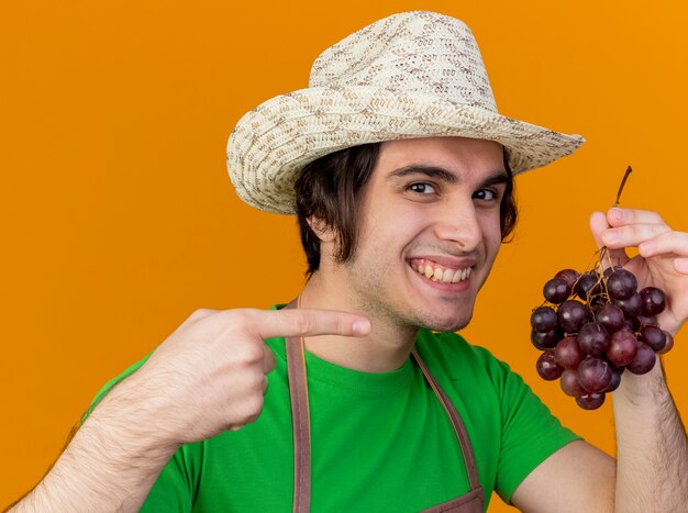 Young gardener man in apron and hat showing bunch of grape pointing with index finger at it smiling with happy face standing over orange wall