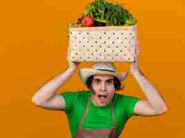 Бесплатное фото Молодой садовник в фартуке и шляпе держит на голове ящик, полный овощей, улыбается со счастливым лицом, стоящим над оранжевой стеной
