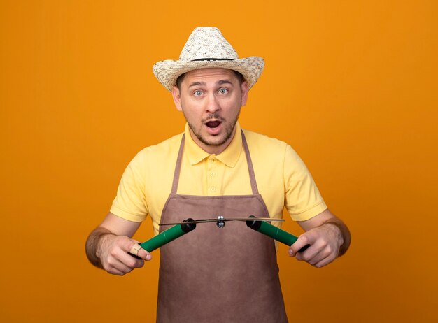 Young gardener man in jumpsuit and hat holding hedge clippers surprised and amazed 