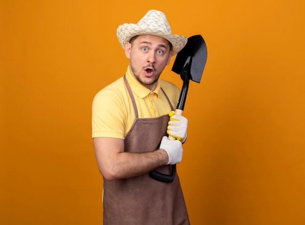 Young gardener man in jumpsuit and hat holding shovel being surprised 