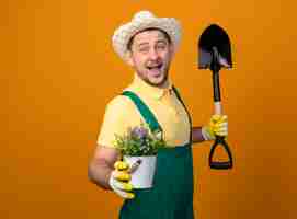 Free photo young gardener man in jumpsuit and hat holding shovel showing potted plant smiling cheerfully