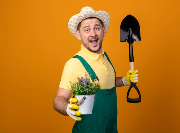 Young gardener man in jumpsuit and hat holding shovel showing potted plant smiling cheerfully 