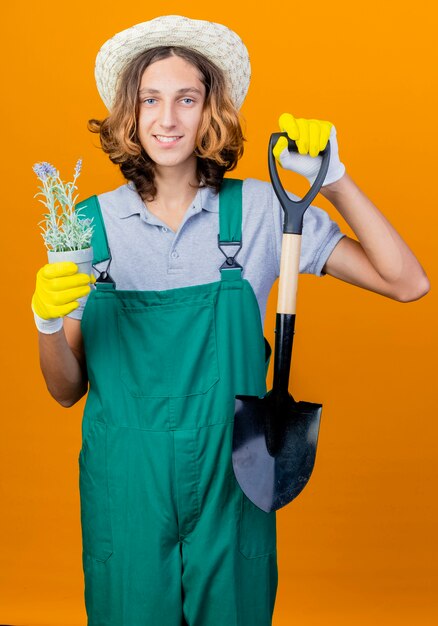 Молодой садовник в резиновых перчатках в комбинезоне и шляпе держит лопату