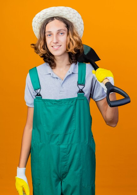 Young gardener man in rubber gloves wearing jumpsuit and hat holding shovel