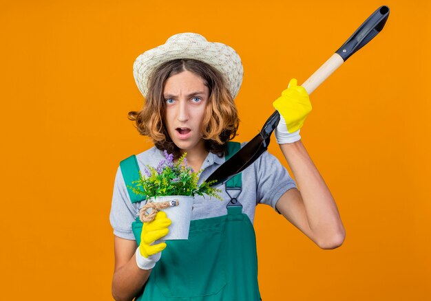 Young gardener man in rubber gloves wearing jumpsuit and hat holding shovel